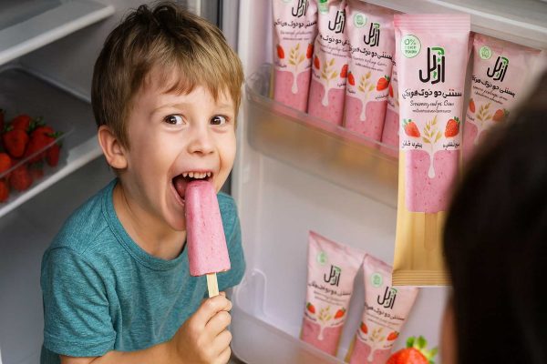 کودک در حال خوردن بستنی گیاهی جو و توتفرنگی آزل جلوی یخچال پر از بستنی و توتفرنگی