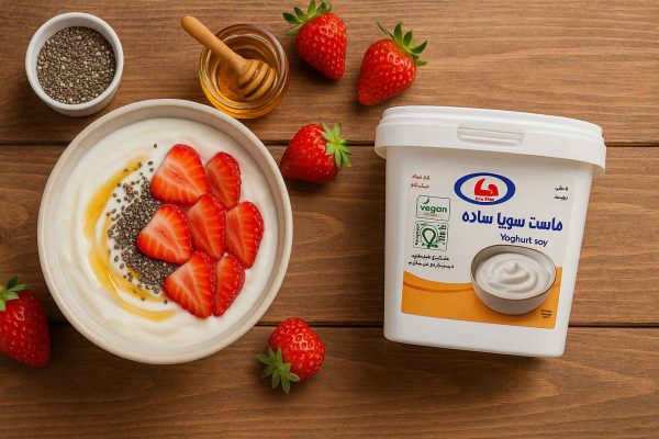 کاسه صبحانه کمقند با ماست سویا ساده دنیا، توتفرنگی تازه، دانه چیا و مقدار کم عسل/اریتریتول؛ نمای از بالا روی میز