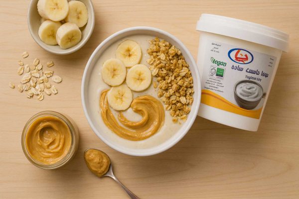 اسموتی پروتئینی گیاهی با ماست سویا ساده دنیا، موز یخزده، اوت/گرنولا و کره بادامزمینی روی میز؛ نمای از بالا