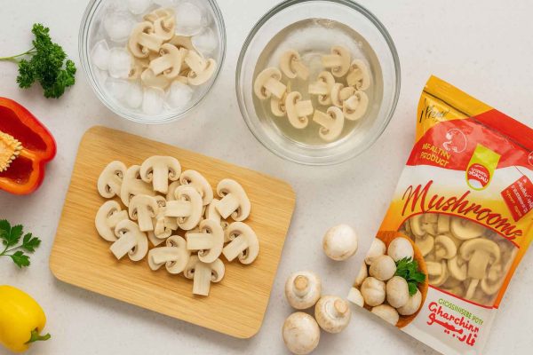 نمای بالا از میز آشپزخانه با قارچ بلنچ قارچینو، قارچهای اسلایسشده، ظرف یخ و سبزیجات تازه