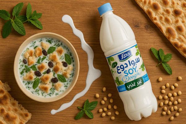 سویا دوغ هراز ۱.۵ لیتری در نمای بالا کنار کاسه آب‌دوغ‌خیار با نان تیلیت‌شده، کشمش و سبزی نعناع، دانه‌های سویا و نان سنگک؛ ریختن تبلیغی دوغ روی میز