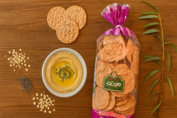 تصویر نان چیپسی جو دوسر و چیا هم‌دانه روی میز چوبی با هوموس، دانه‌های چیا و جو دوسر و شاخه بید مجنون | سورن فودز