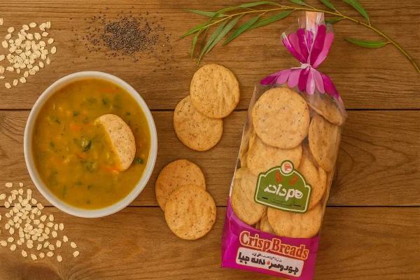 تصویر نان چیپسی جو دوسر و چیا هم‌دانه روی میز چوبی بیدی با ظرف سوپ، دانه‌های چیا و جو دوسر و شاخه بید مجنون | سورن فودز