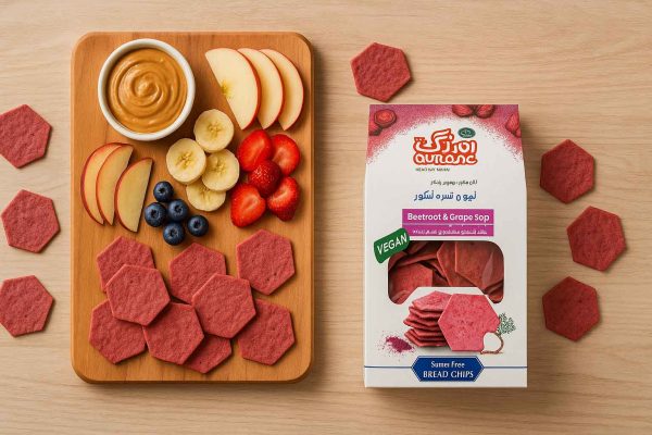 نما از بالا روی میز چوب سپیدار؛ برد صبحانه سبک با برش میوه و کره بادام‌زمینی کنار نان چیپس لبو و شیره انگور اورنگ، همراه با جعبه محصول