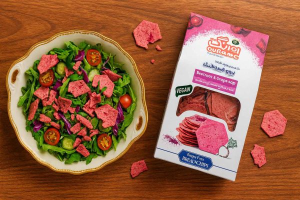 نما از بالا روی میز چوب نارون؛ سالاد کنار غذا با تکه‌های نان چیپس لبو و شیره انگور اورنگ به‌جای کروتون، همراه با جعبه محصول